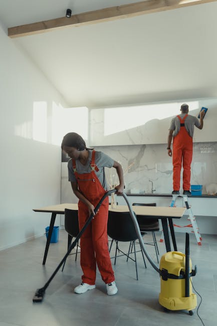 Two professional cleaning workers from Bayswater Carpet Cleaners are performing deep surface cleaning in a modern, well-lit kitchen. The worker in the foreground is using a vacuum or carpet cleaner on